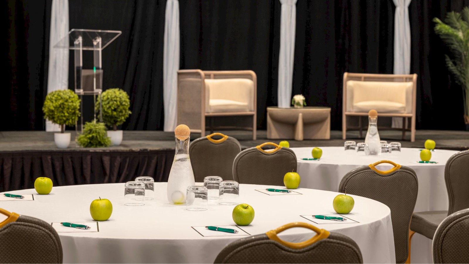 Conference room with round tables, green apples, water bottles, and glasses; a stage with chairs and a podium in the background.