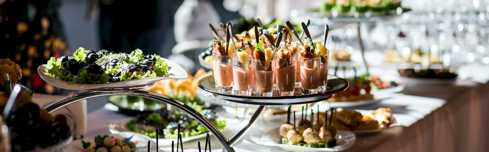 A buffet table with a variety of appetizers, pastries, and drinks set on different tiers under bright lighting.