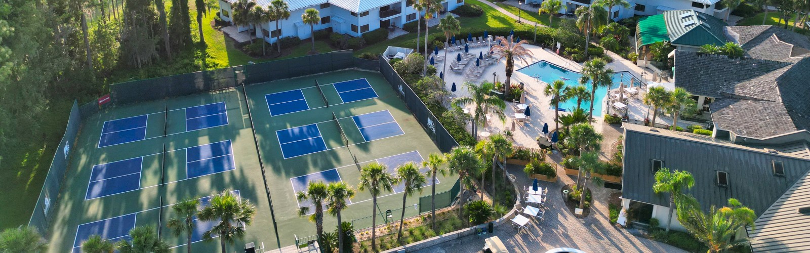 Aerial view of a resort with tennis courts, a pool area, white-roofed buildings, and surrounding greenery.