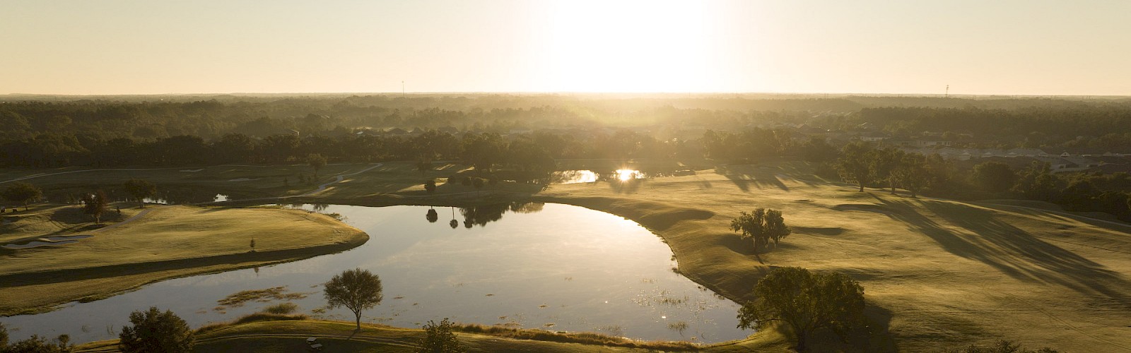 Sunset over a golf course with winding water hazards, green fairways, and a few white-roofed buildings in the foreground.