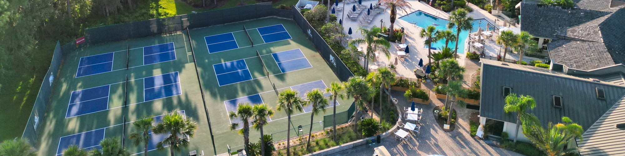 Aerial view of a resort with tennis courts, a pool area, white-roofed buildings, and surrounding greenery.