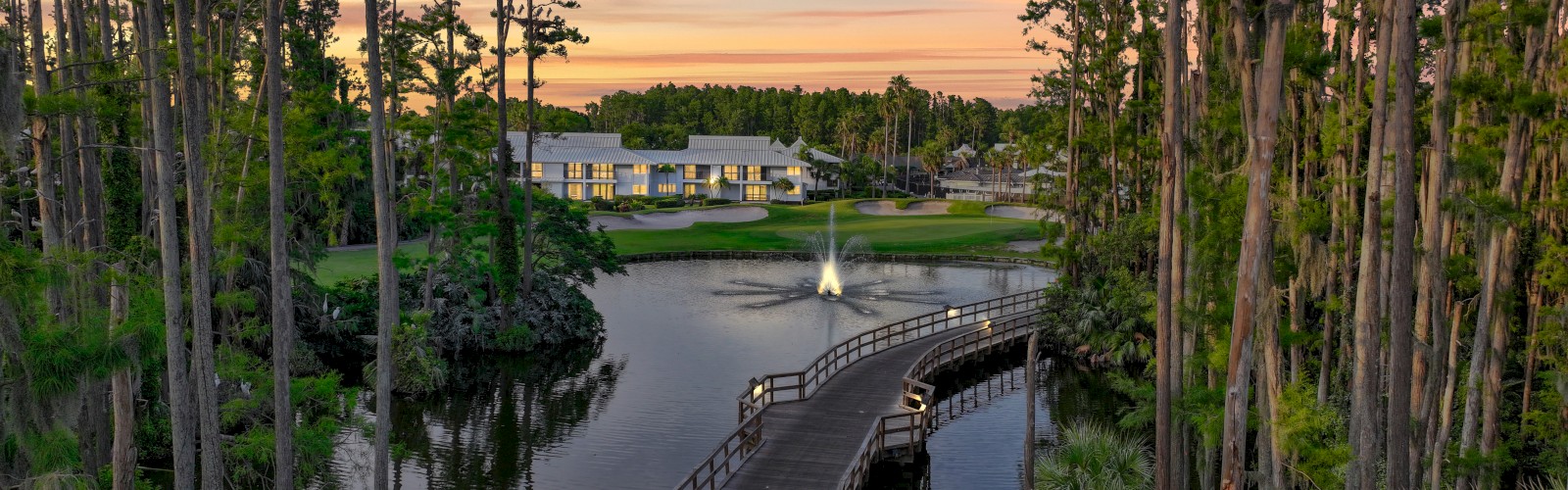 A scenic view of a wooden bridge over a tranquil lake, surrounded by tall trees, leading to a building, with a fountain in the water.