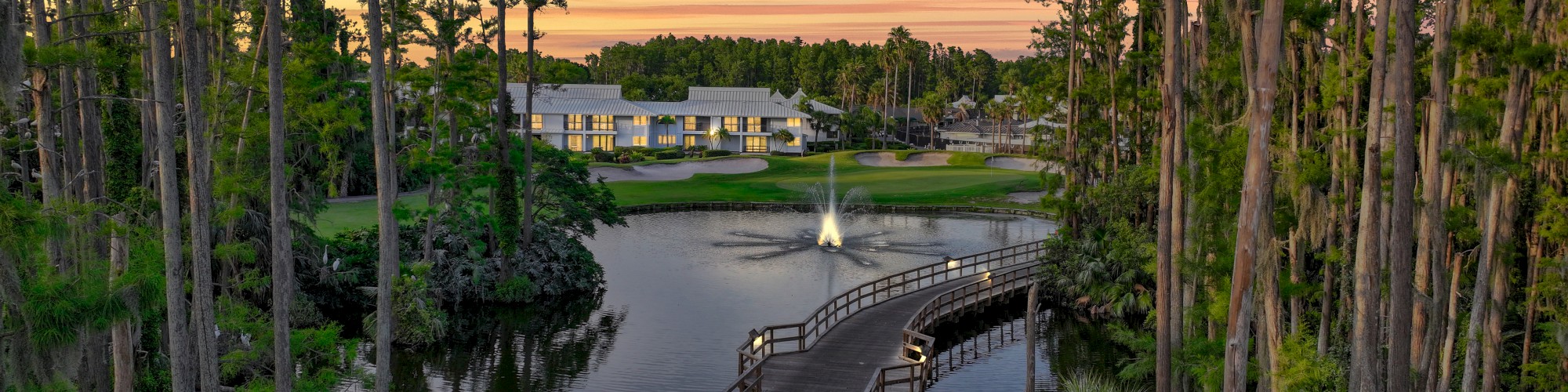 A scenic view of a wooden bridge over a tranquil lake, surrounded by tall trees, leading to a building, with a fountain in the water.