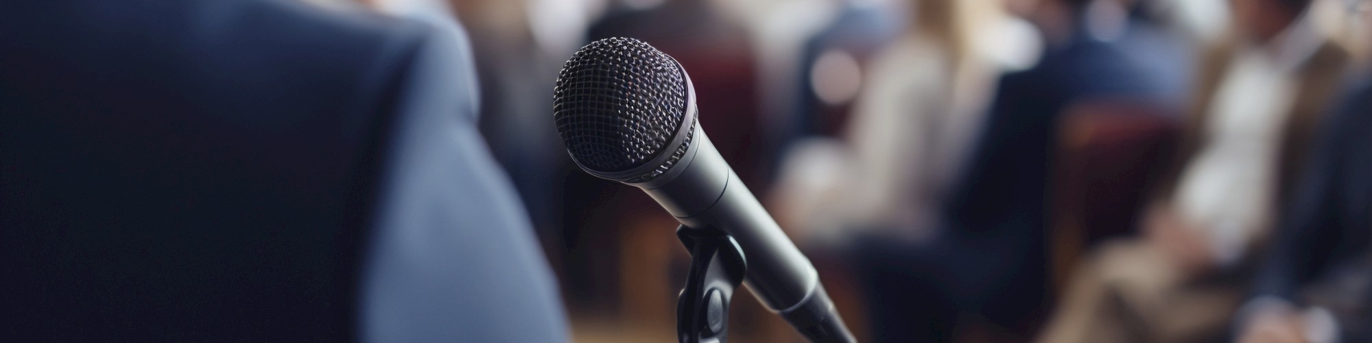 A person standing near a microphone in front of an audience, suggesting a public speaking or presentation event.