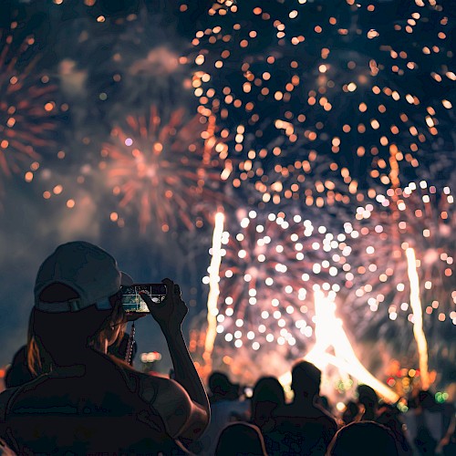 A person wearing a cap takes a photo of a fireworks display with a crowd watching in the foreground at night.