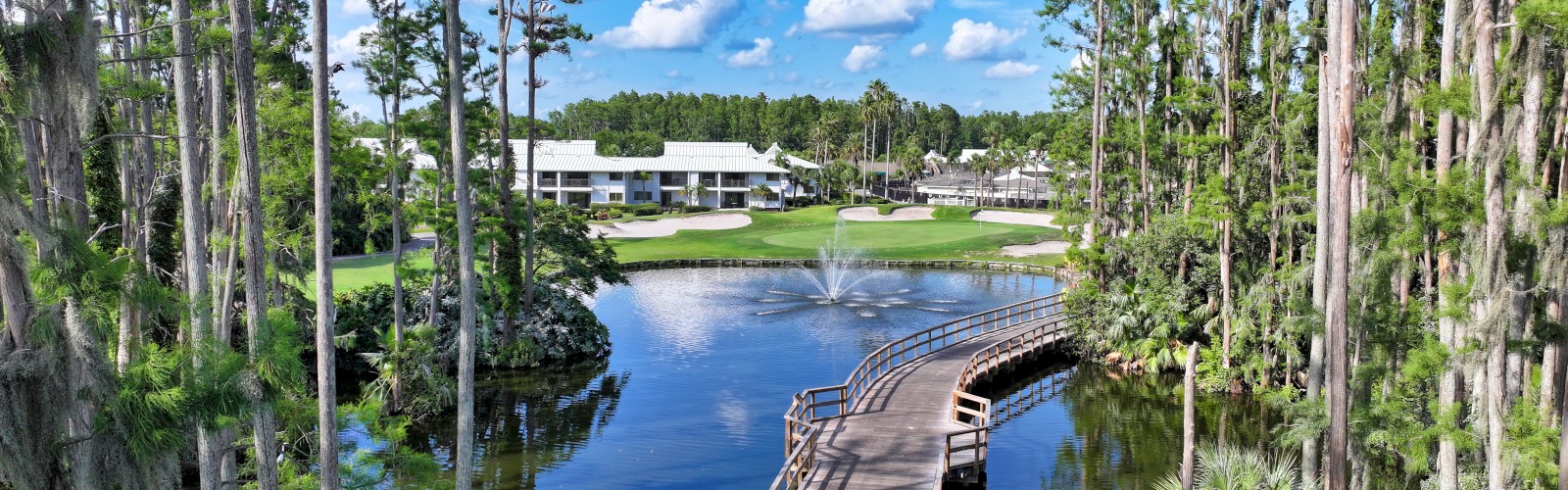 A wooden path spans a tranquil lake surrounded by tall trees, leading to a building in the distance. A fountain sprays in the lake.