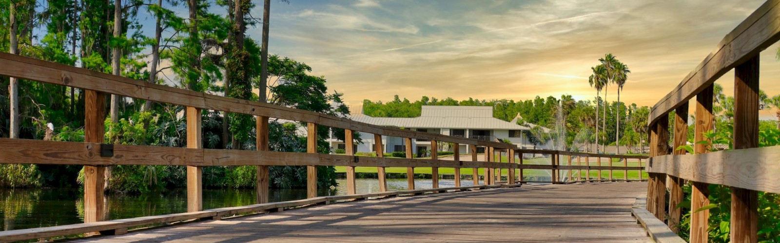 A wooden walkway leads through a tranquil park, flanked by tall trees, with a serene sky and distant buildings in the background.