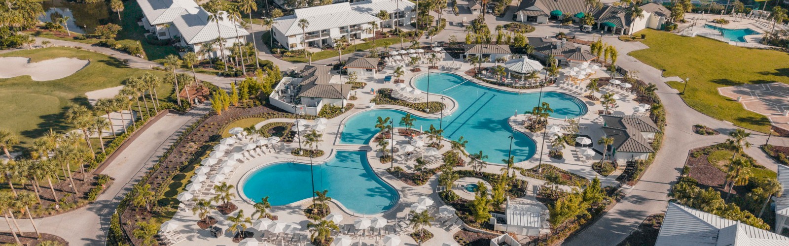 An aerial view of a resort with swimming pools, lounge chairs, palm trees, surrounded by residential buildings and pathways.