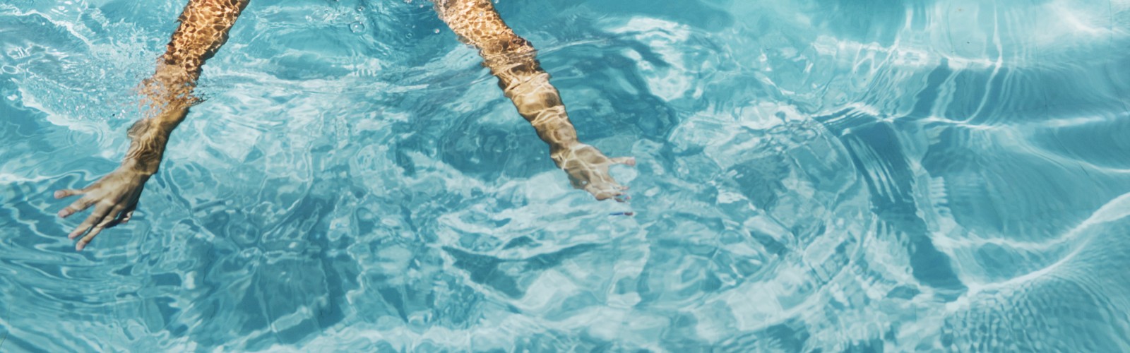A pair of legs submerged in a swimming pool with clear blue water and ripples forming around them, under sunlight, creating a serene view.