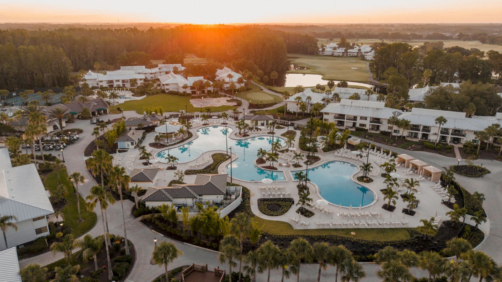 Sunset over a luxurious resort with multiple pools, palm trees, white buildings, and a golf course in the background.
