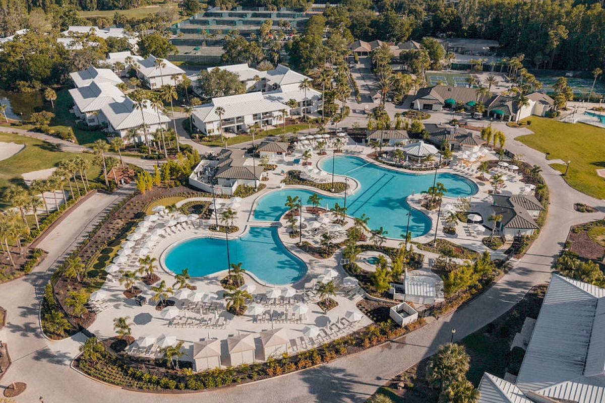 Aerial view of a luxurious resort with multiple pools, lounge chairs, palm trees, and surrounding buildings in a lush, green landscape.