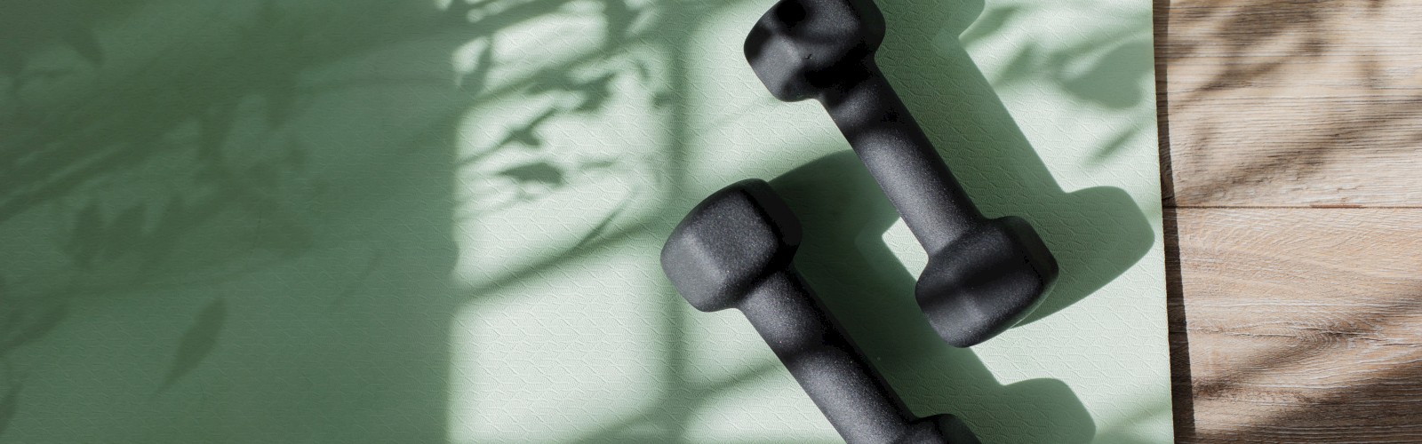 Two black dumbbells on a green yoga mat with shadows of leaves, creating a peaceful workout ambiance.
