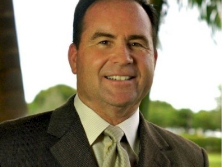A smiling man in a suit outdoors, with trees and greenery in the background, on a bright day.