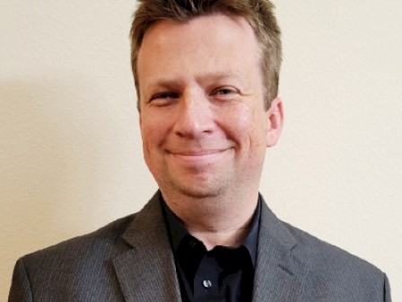 A smiling man in a dark suit and shirt stands against a plain light-colored wall.