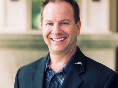 A smiling man in a suit, standing outdoors in front of a building with columns, wearing a lapel pin that resembles a waving flag.