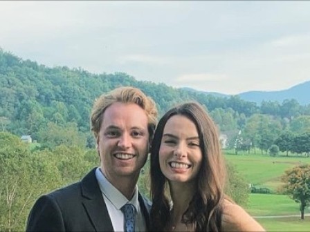 A man and woman are smiling outdoors with a scenic background of green trees, rolling hills, and mountains in the distance.