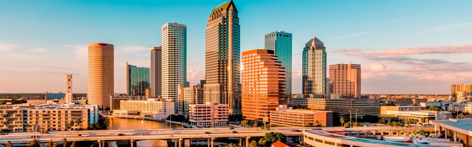 A scenic view of a cityscape with tall buildings, a river, and a bridge under a clear blue sky, illuminated by a setting sun.