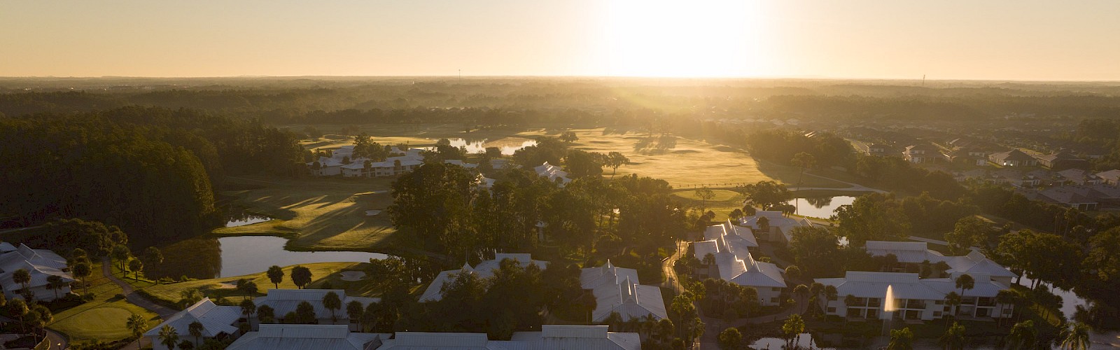 An aerial view of a resort complex at sunset, featuring a large pool area, courtyards, palm trees, and low buildings.
