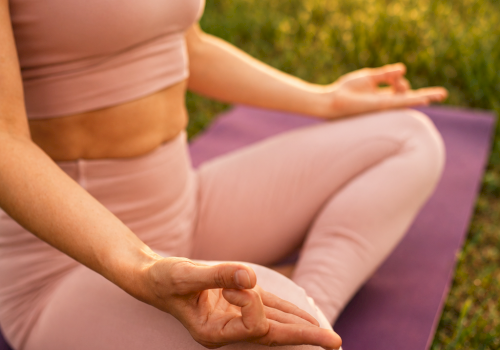 A woman is practicing yoga outdoors during sunset, sitting cross-legged on a purple mat, with trees and sunshine in the background.