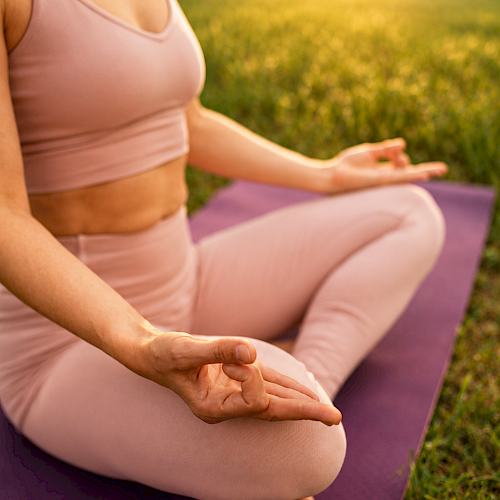 A woman is practicing yoga outdoors during sunset, sitting cross-legged on a purple mat, with trees and sunshine in the background.