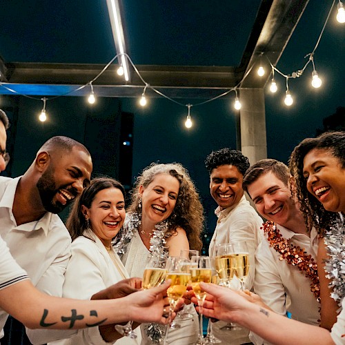 A diverse group of friends celebrating together with drinks at an outdoor party under string lights at night.