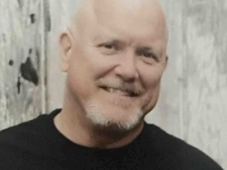 A smiling bald man with a goatee stands in front of a weathered wooden background.