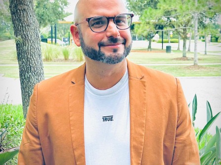 A man with glasses and a beard is smiling outdoors, wearing an orange blazer and a white T-shirt with 