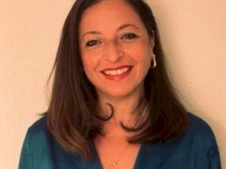 A woman with straight brown hair, smiling, wearing a blue top and a gold necklace, standing against a beige wall.