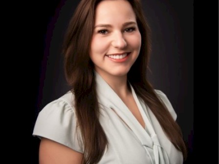 A smiling young woman with long brown hair, wearing a light-colored blouse, against a dark background.