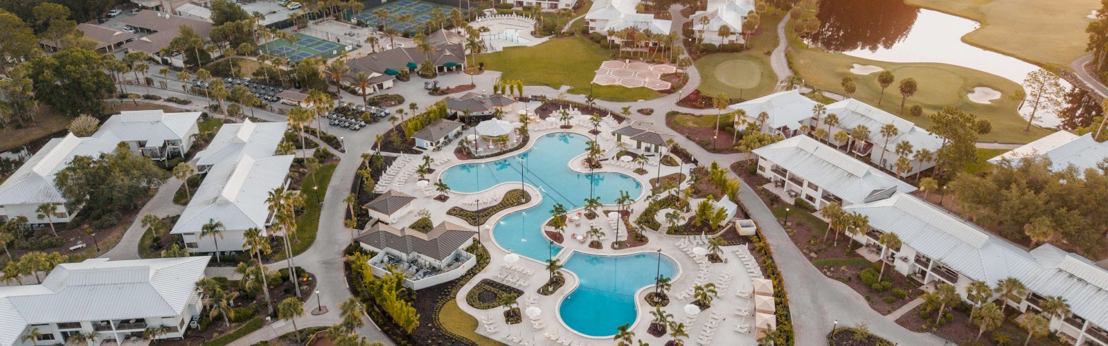 An aerial view of a resort with swimming pools, white-roofed buildings, tennis courts, a golf course, and lush greenery at sunset.