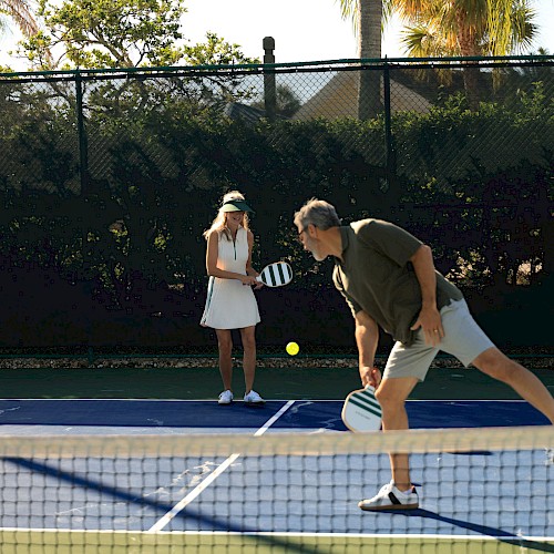 A man and woman are playing pickleball on a court with a net, surrounded by trees and a fence, on a sunny day.