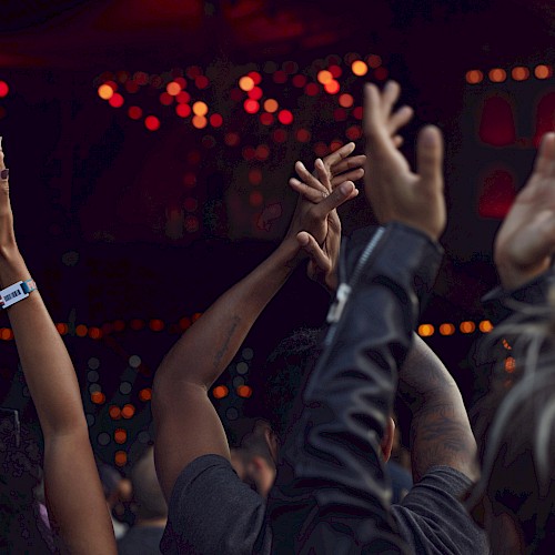 People raising their hands in the air at a concert or event, with vibrant red and orange lights in the background.