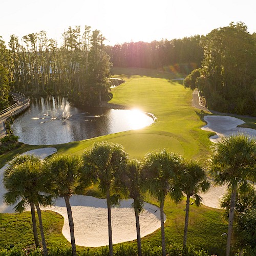 A golf course with sand traps, water hazards, and palm trees is illuminated by the setting sun.