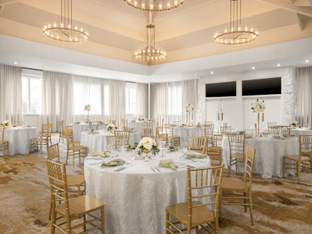 A beautifully decorated banquet hall with round tables covered in white cloths, elegant gold chairs, and floral centerpieces.