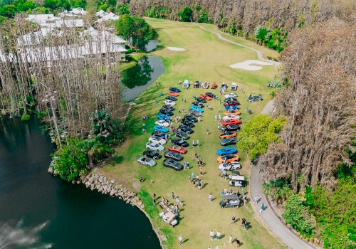 A golf course event with colorful cars, golf carts, and people enjoying a sunny day near water and trees.