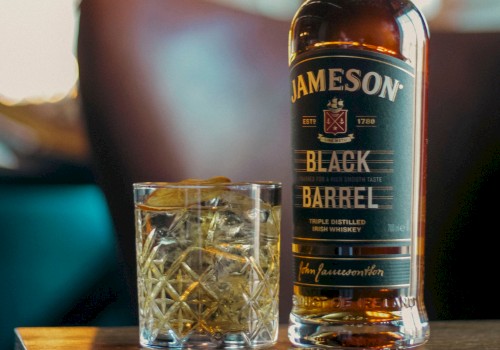 A bottle of Jameson Black Barrel Irish whiskey and a glass with ice and lemon on a wooden table in a cozy bar setting.