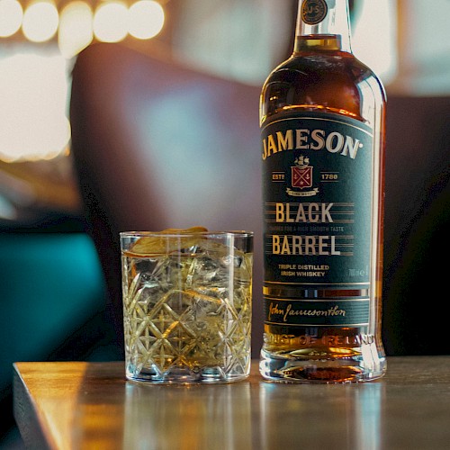 A bottle of Jameson Black Barrel Irish whiskey and a glass with ice and lemon on a wooden table in a cozy bar setting.