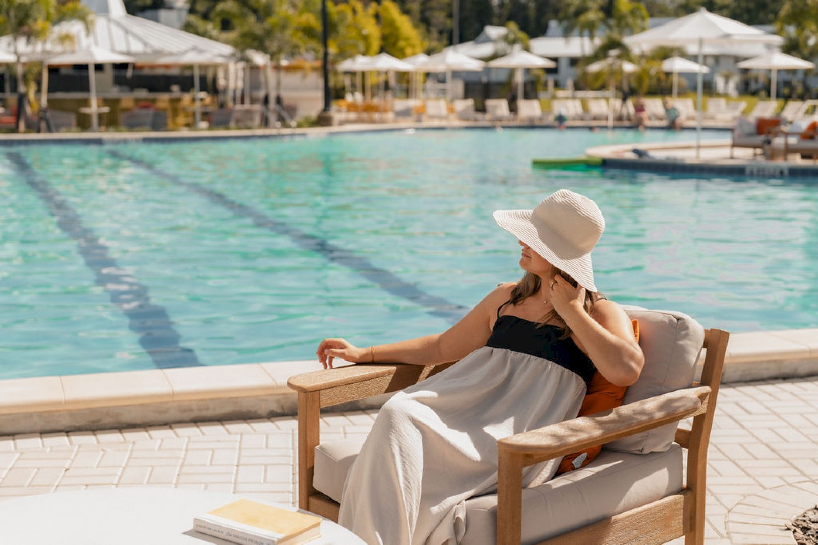 A woman relaxes by the pool, sitting on a cushioned chair, wearing a large sun hat and summery dress on a sunny day.