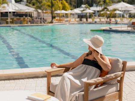 A woman relaxes by the poolside on a sunny day, sitting on a cushioned chair with a large, floppy hat shielding her face.