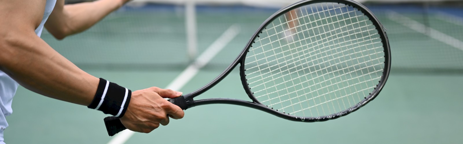 A person prepares to serve a tennis ball on a court, holding a racket, with a net and opponent visible in the background.