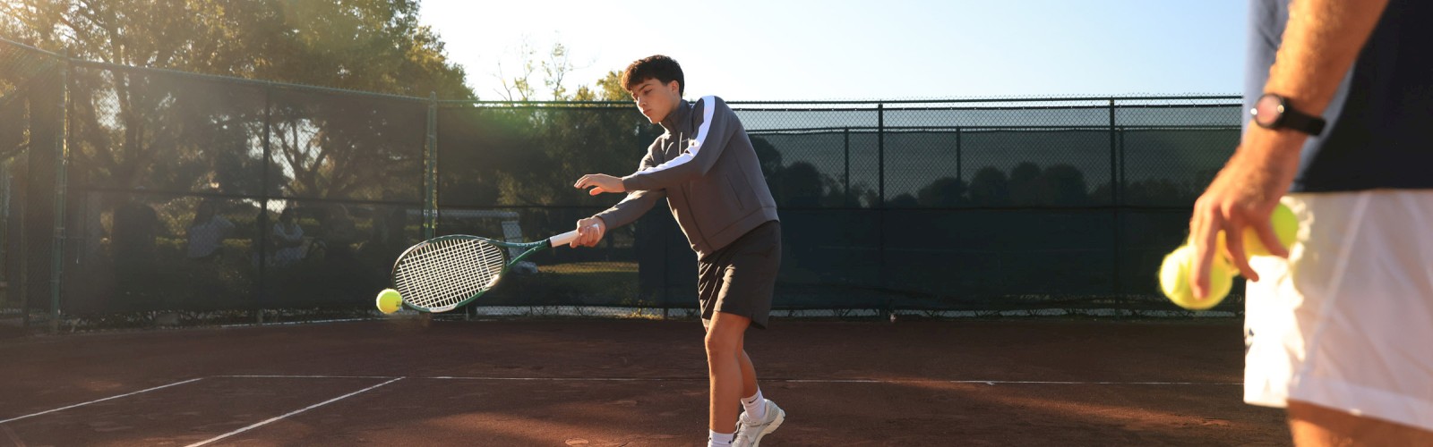 A tennis player lunges to return a serve on a clay court as another player watches, the sun low on the horizon and shadows stretching long.