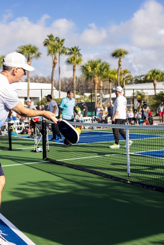 People are playing pickleball on outdoor courts surrounded by palm trees, with spectators and other players in the background.