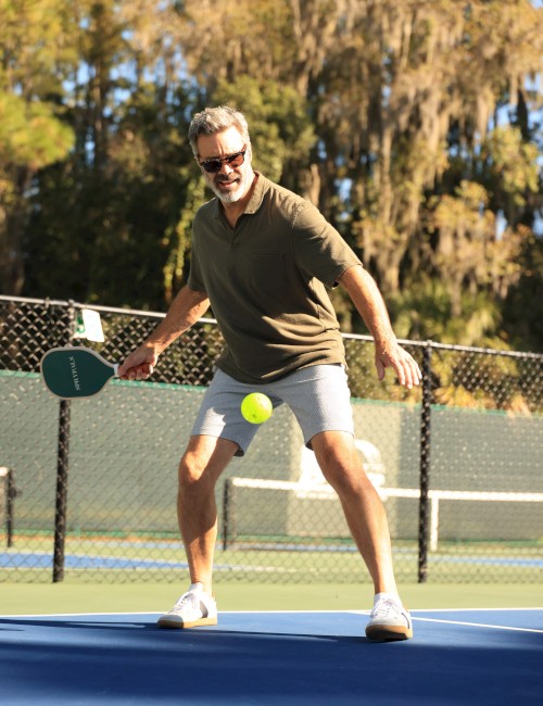 A man in a green shirt and gray shorts plays tennis on a blue court, ready to hit the ball, with a fence and trees in the background.