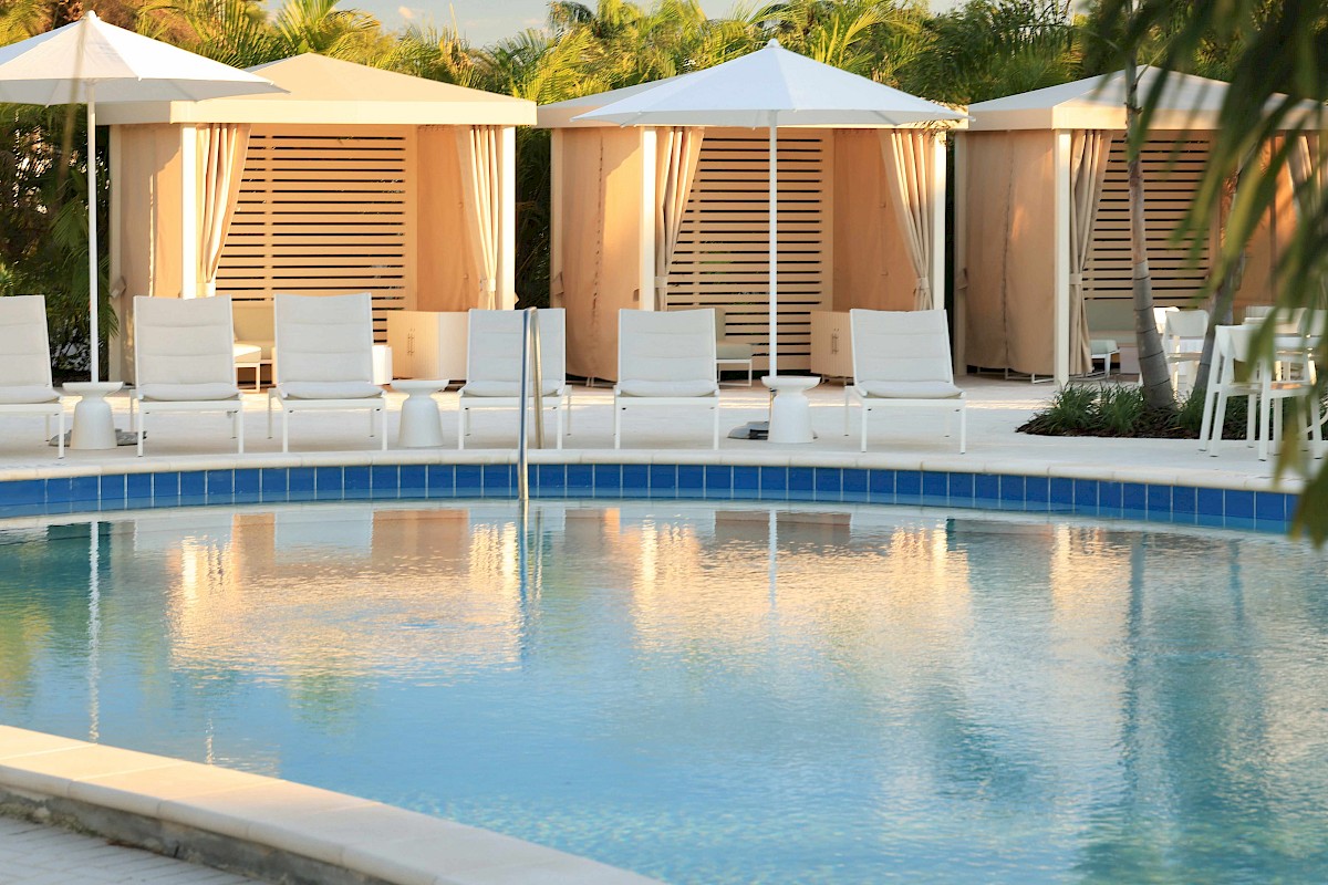 The image shows a serene swimming pool area with white lounge chairs, umbrellas, and private cabanas surrounded by lush greenery.