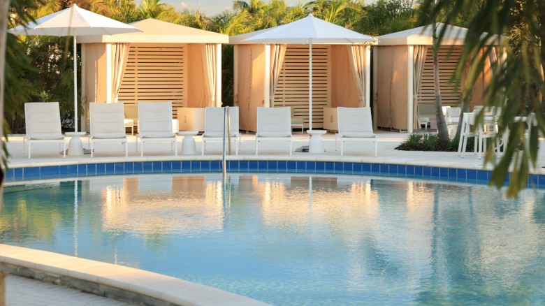 The image shows a serene swimming pool area with white lounge chairs, umbrellas, and private cabanas surrounded by lush greenery.