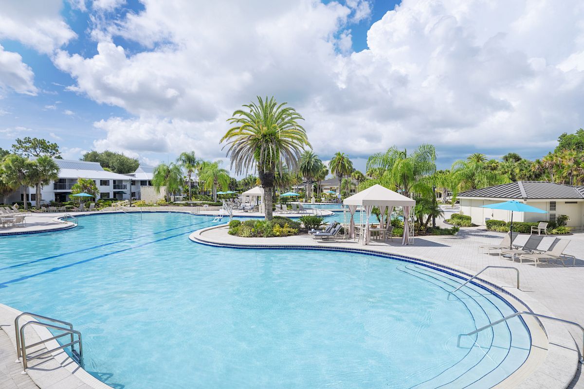 An outdoor swimming pool area with lounge chairs, palm trees, and cabanas. The sky is partly cloudy, and there are buildings in the background.