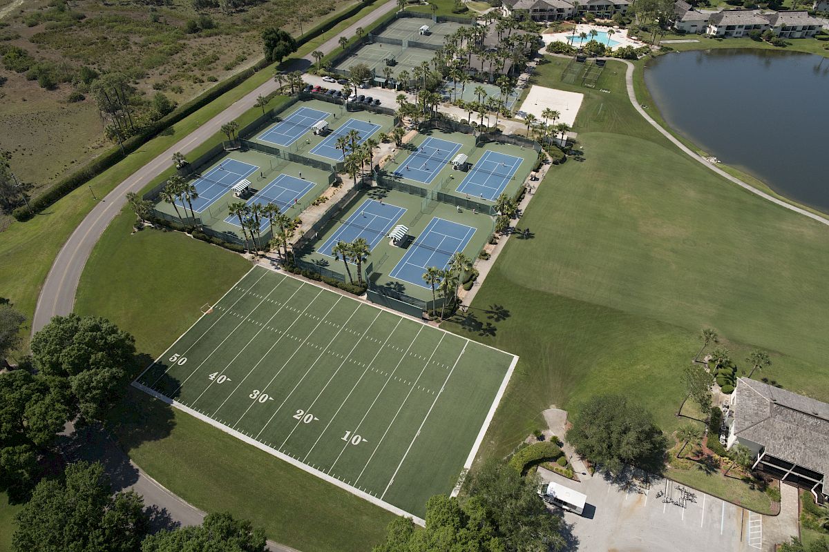 Aerial view of a sports complex with six tennis courts, a small football field, a lake, and nearby buildings under clear skies, ending the sentence.