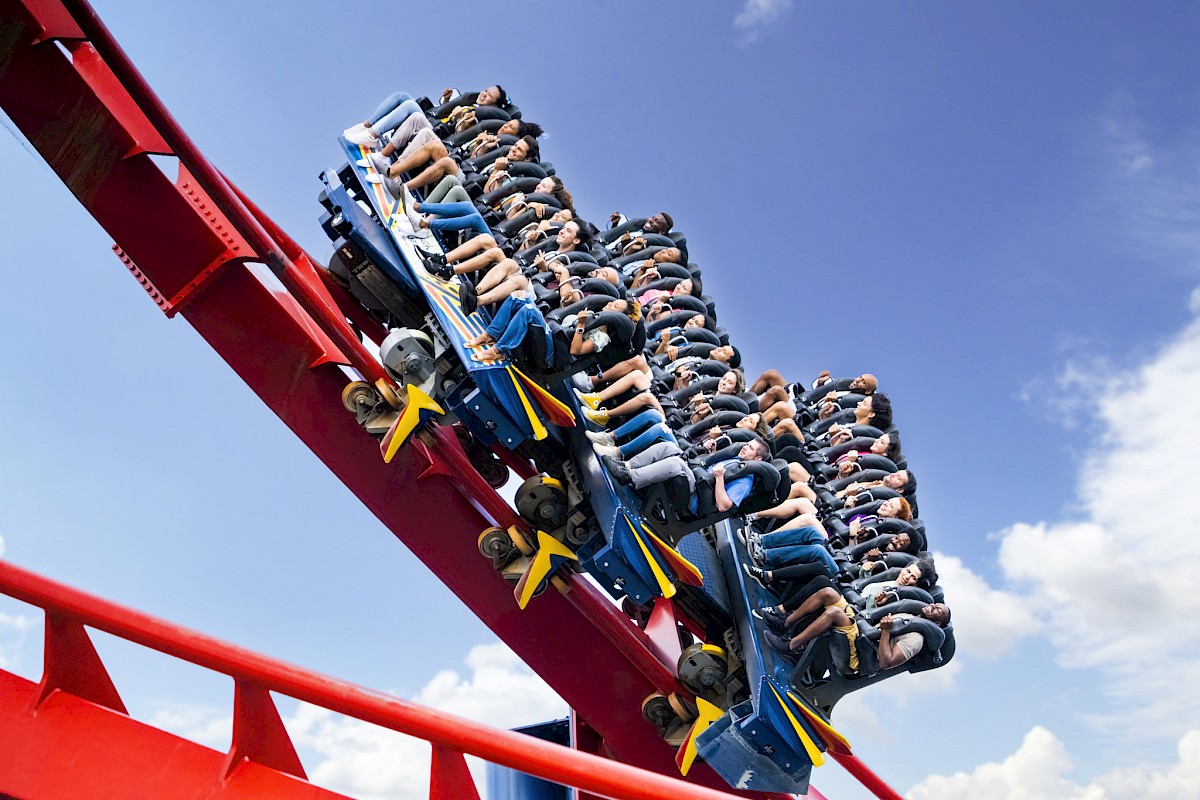 A roller coaster with people riding, faces blurred in motion, on a steep red track against a bright blue sky with scattered clouds.