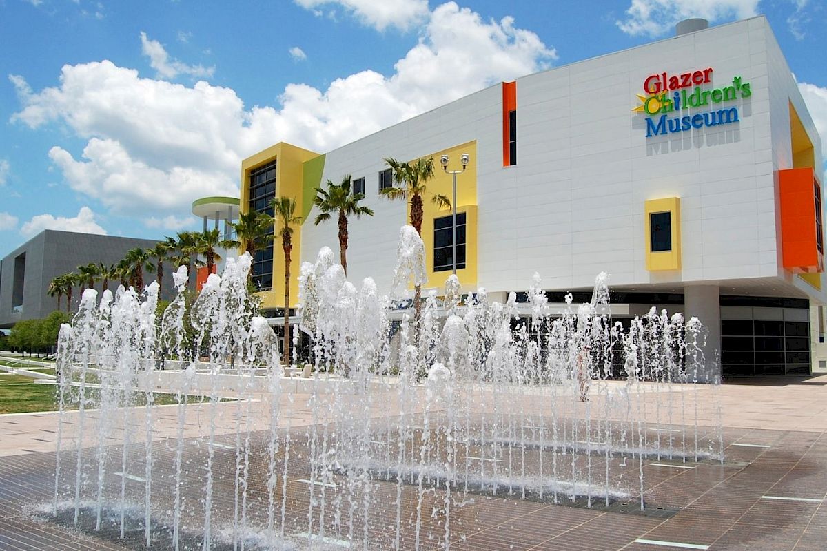 The image shows the exterior of Glazer Children's Museum with a splash fountain in the foreground, set against a bright, partly cloudy sky.