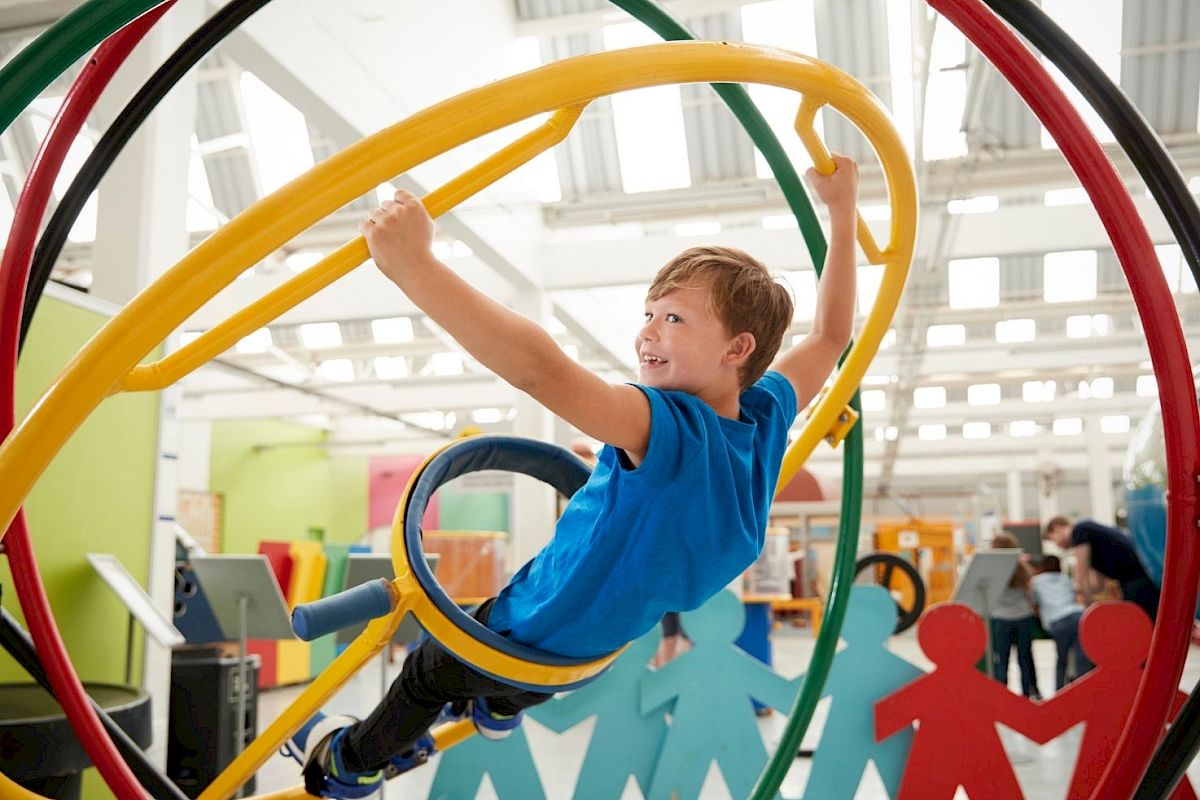 A smiling child is playing on a colorful climbing structure indoors, surrounded by large cutouts of people holding hands.